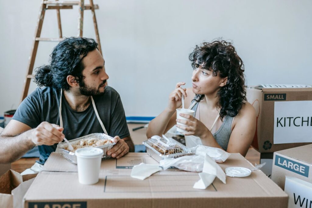 Two people eating takeout meals among moving boxes, illustrating tips for staying healthy while you’re between homes.