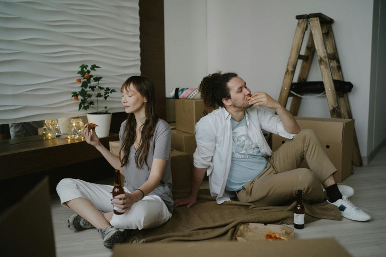 Couple sitting on the floor eating and drinking beer during a move, surrounded by boxes.