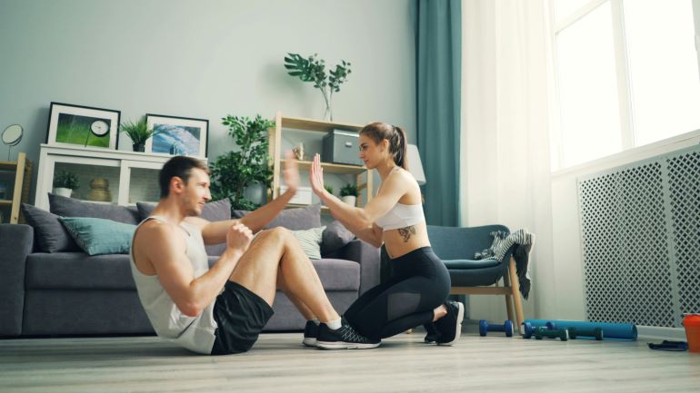 Two people exercising on a living room floor with home workout gear nearby, practicing movement and support together.