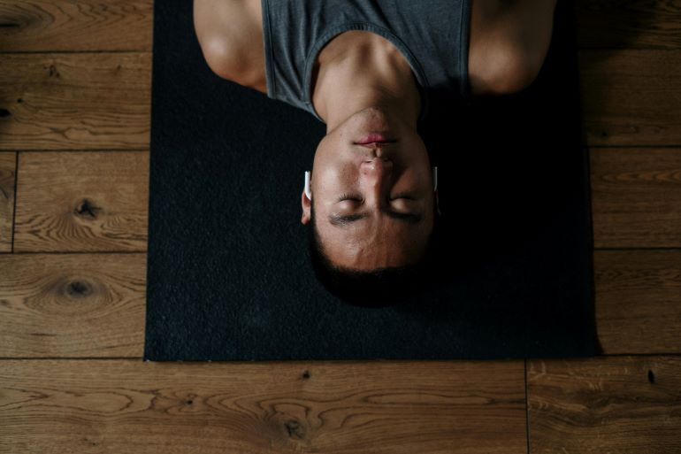Tag: Person lying on a yoga mat with eyes closed, resting on a wooden floor during a relaxing recovery moment.